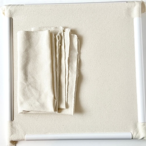 May include: A white canvas stretched over a white wooden frame. The canvas is textured and has a slight sheen. There are several pieces of white fabric on top of the canvas.