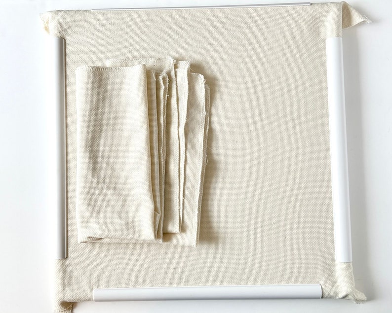 May include: A white canvas stretched over a white wooden frame. The canvas is textured and has a slight sheen. There are several pieces of white fabric on top of the canvas.