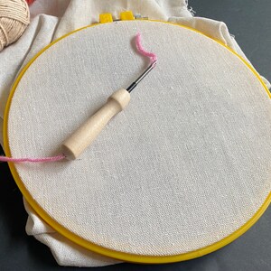 Off white cotton linen fabric stretched over an 8 inch plastic yellow embroidery hoop with a 3mm punch needle with wood handle sitting on it for punch needle or cross stitch work.
