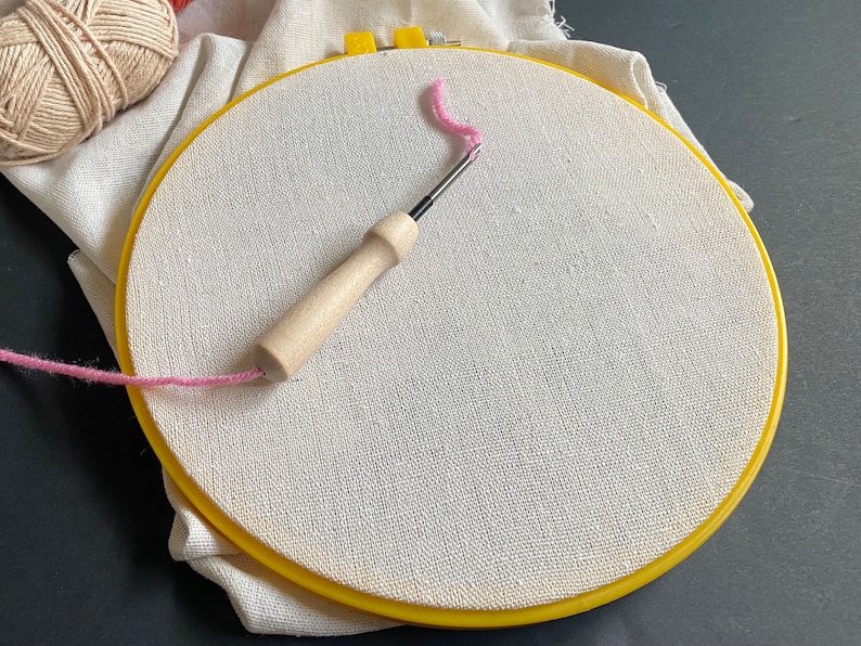 Off white cotton linen fabric stretched over an 8 inch plastic yellow embroidery hoop with a 3mm punch needle with wood handle sitting on it for punch needle or cross stitch work.