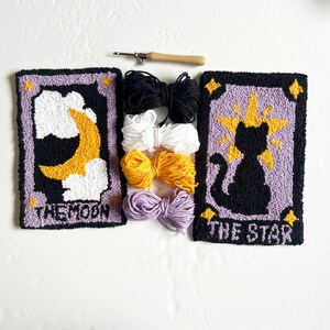 May include: Two punch needle embroidery kits featuring the tarot cards The Moon and The Star. The Moon card features a yellow crescent moon and clouds on a purple background. The Star card features a black cat silhouette with a yellow star on a purple background. Both kits include embroidery floss in black, white, yellow, and purple.