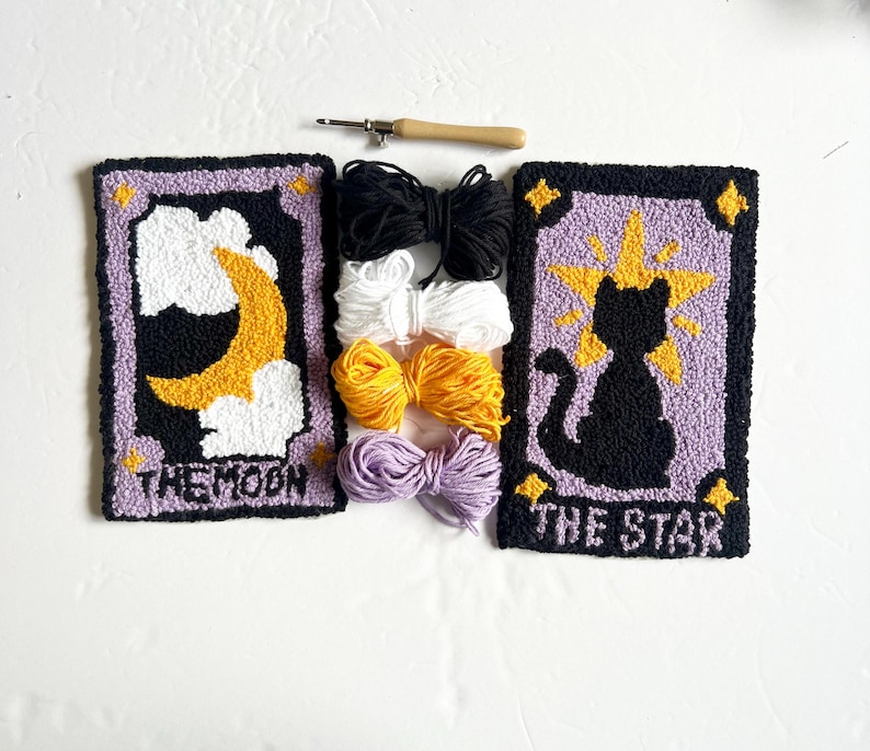May include: Two punch needle embroidery kits featuring the tarot cards The Moon and The Star. The Moon card features a yellow crescent moon and clouds on a purple background. The Star card features a black cat silhouette with a yellow star on a purple background. Both kits include embroidery floss in black, white, yellow, and purple.