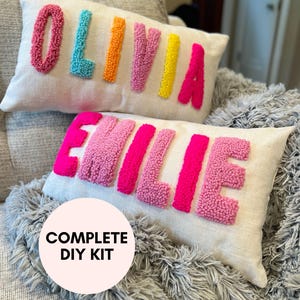 May include: Two white pillow covers with the names "Olivia" and "Eillie" spelled out in colorful yarn pom-poms. The pillows are on a gray fuzzy blanket. A white circle with the text "COMPLETE DIY KIT" is in the foreground.