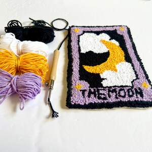 May include: A punch needle kit featuring a purple and black card with a yellow crescent moon and white clouds. The card reads "THE MOON". The kit includes four balls of yarn in purple, yellow, white, and black, and a wooden punch needle.