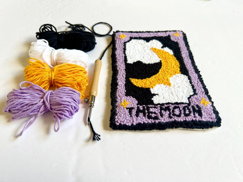 May include: A punch needle kit featuring a purple and black card with a yellow crescent moon and white clouds. The card reads "THE MOON". The kit includes four balls of yarn in purple, yellow, white, and black, and a wooden punch needle.