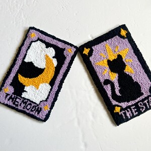 May include: Two purple and black tarot cards with embroidered designs. The card on the left is "The Moon" and features a yellow crescent moon in white clouds. The card on the right is "The Star" and features a black cat with a yellow starburst behind it.