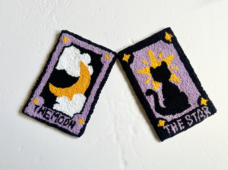May include: Two purple and black tarot cards with embroidered designs. The card on the left is "The Moon" and features a yellow crescent moon in white clouds. The card on the right is "The Star" and features a black cat with a yellow starburst behind it.
