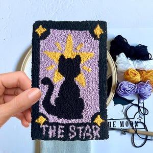 May include: A punch needle kit featuring a black cat with a yellow star above it. The card reads "The Star". The kit includes yarn in black, white, yellow, and purple.