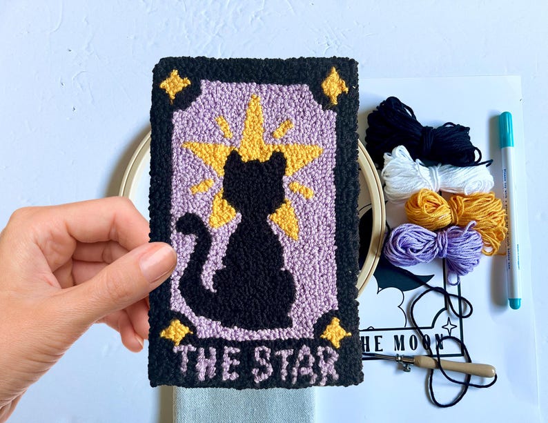 May include: A punch needle kit featuring a black cat with a yellow star above it. The card reads "The Star". The kit includes yarn in black, white, yellow, and purple.