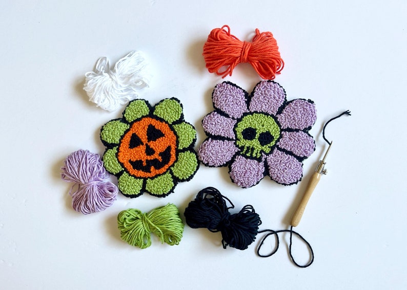 mug rug punch needle coasters. one is an orange jack o lantern face with green flower petals, the other is a green skull with purple flower petals. they are surrounded by purple orange white green black yarn and a punch needle.