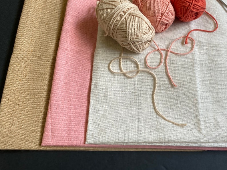 3 pieces of cotton linen fabric for punch needle or needlepoint in khaki brown, pink, and off white with three mini skeins of cotton yarn in beige, pink, and burnt orange on top of them.