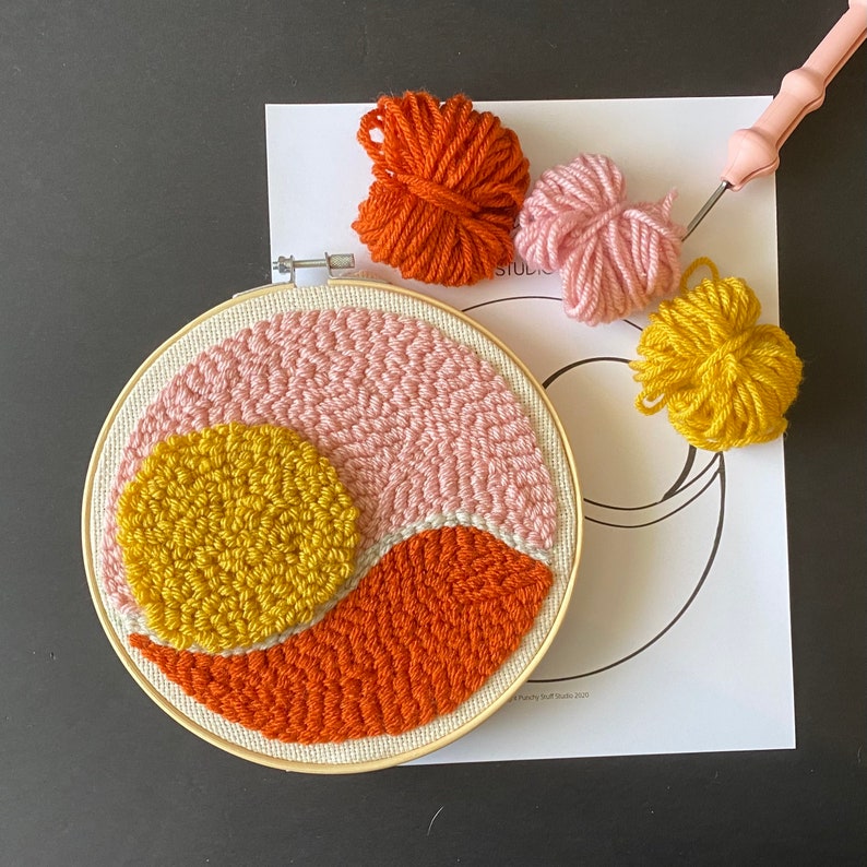May include: A close-up of a colorful embroidery hoop with a pink, orange, and yellow design. The hoop is on a white paper with a black and white outline of a crescent moon. There are three balls of yarn in the background, one pink, one orange, and one yellow.