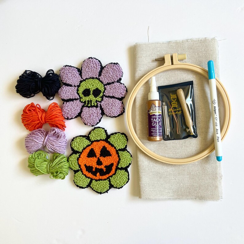 two mug rugs one with a green skull in the center with purple flower petals, the other with a orange jack o lantern pumpkin in the middle and green petals, 4 skeins of yarn in black orange purple and green. Linen fabric, a fabric pen, glue, hoop