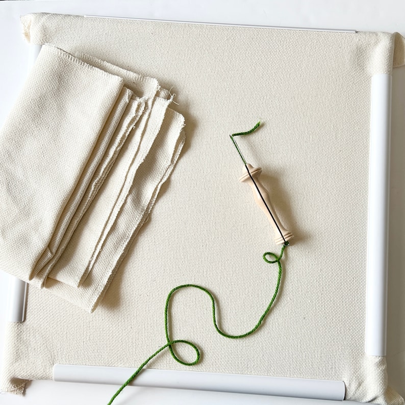 May include: A white embroidery hoop with a wooden needle threader and green embroidery floss. A stack of white fabric is to the left of the hoop.