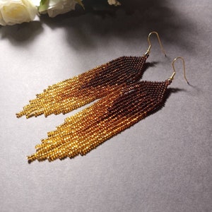 May include: A pair of long, beaded earrings with a gradient of brown and gold beads. The earrings are made with small, round beads that are strung together to create a fringe effect. The earrings have gold hooks.