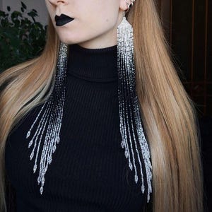 Puede incluir: Pendientes largos colgantes con diseño de cuentas. Los pendientes presentan un degradado de cuentas negras a plateadas, creando un efecto visual llamativo. Se llevan con un cuello alto negro.