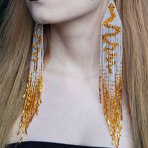 May include: Long, beaded earrings with a gray and gold design. The earrings feature a zigzag pattern in gold beads against a gray background, with a gold fringe at the bottom. The earrings are a statement accessory.