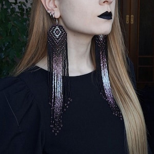 Puede incluir: Una mujer con el pelo largo y rubio que lleva ropa negra y pendientes largos de cuentas con un patrón geométrico. Los pendientes son negros, blancos y morados.