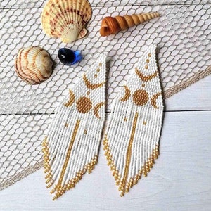 Può includere: Orecchini con perline bianche e dorate con un design a frangia. Gli orecchini presentano un motivo di luna crescente e sole.