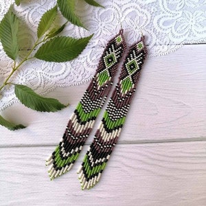 May include: A pair of long, beaded earrings with a geometric pattern in shades of green, white, and purple. The earrings are hanging from a white surface.