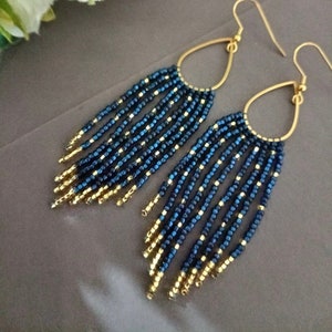 Peut inclure: Une paire de longues boucles d'oreilles pendantes avec des perles bleues et dorées. Les boucles d'oreilles sont faites de petites perles rondes enfilées dans un motif en cascade. Les perles sont d'un bleu profond avec un reflet métallique, et elles sont entrecoupées de perles dorées. Les boucles d'oreilles sont attachées à des anneaux dorés.