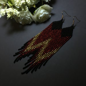 May include: A pair of long, beaded earrings with a black, red and gold colour scheme. The earrings are made of small beads that are strung together to create a fringe effect.