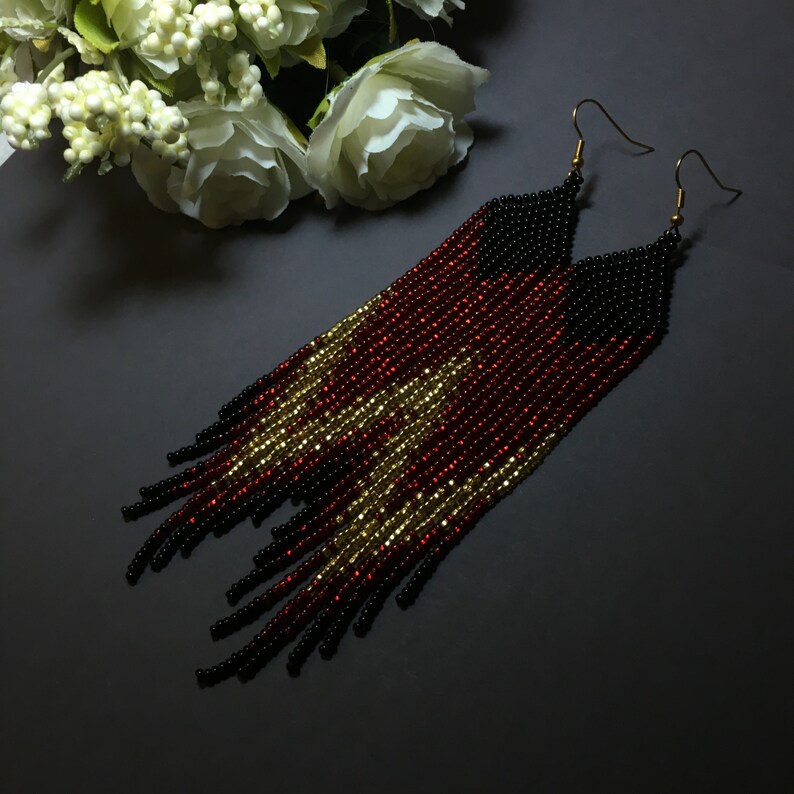 May include: A pair of long, beaded earrings with a black, red and gold colour scheme. The earrings are made of small beads that are strung together to create a fringe effect.