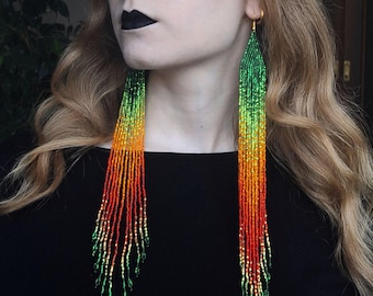 May include: Long, colorful beaded earrings with a gradient of green, orange, and red beads. The earrings are hanging from the ears of a person with long, wavy hair.