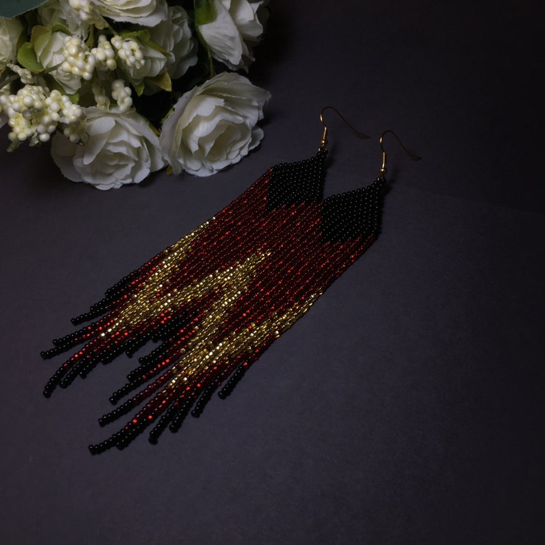 May include: A pair of long, beaded earrings with a black, red and gold colour scheme. The earrings are made of small beads and have a fringe-like design.