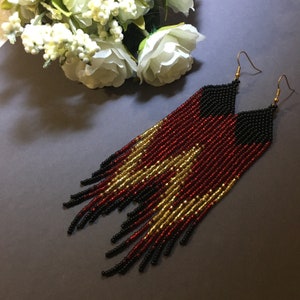 May include: A pair of long, beaded earrings with black, red and gold beads. The earrings are made with a fringe design and have a gold hook fastening.