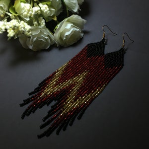 May include: Long beaded earrings with a black, red and gold colour scheme. The earrings are made of small beads and have a fringe design.