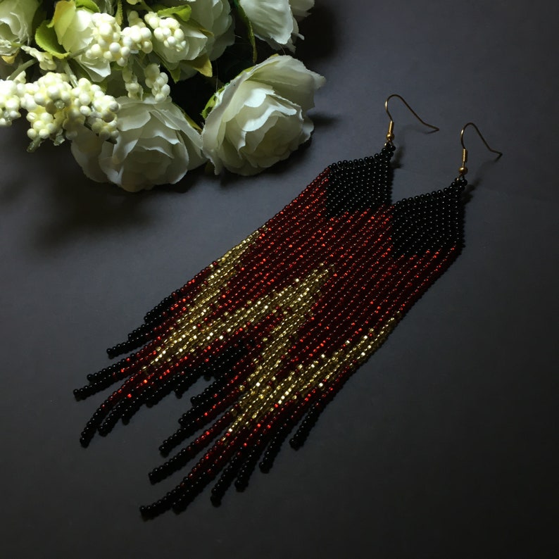 May include: Long beaded earrings with a black, red and gold colour scheme. The earrings are made of small beads and have a fringe design.