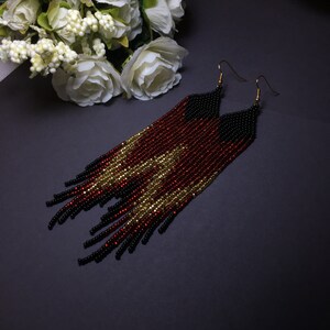 May include: A pair of long, beaded earrings with a black, red and gold colour scheme. The earrings are made of small beads and have a fringe design.