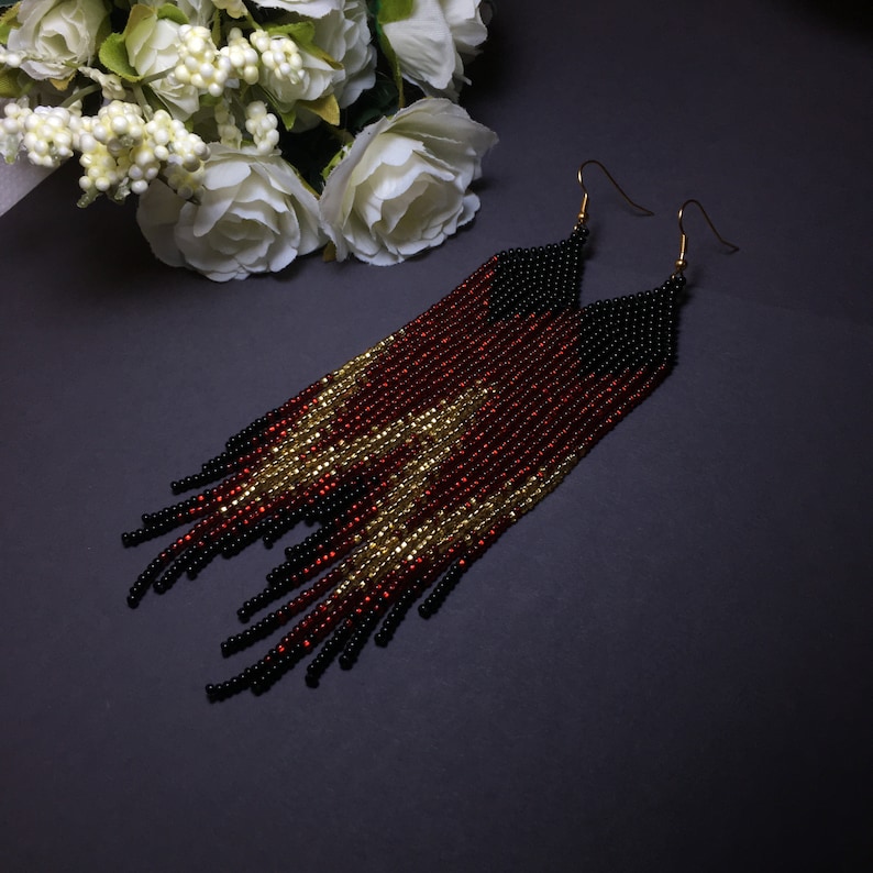 May include: A pair of long, beaded earrings with a black, red and gold colour scheme. The earrings are made of small beads and have a fringe design.