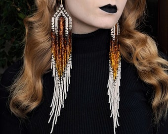 May include: Long, beaded earrings with a white and brown ombre design. The earrings are made of small, round beads and have a geometric pattern at the top.