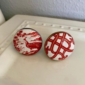 May include: Two round ceramic drawer knobs with a white background and red designs. One knob features a red bird design and the other has a red geometric pattern.