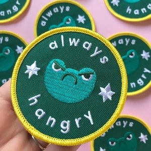 Puede incluir: Parche bordado verde y amarillo con una cara de rana de dibujos animados y el texto "always hangry" en blanco.