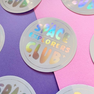 May include: Circular holographic stickers with the text "SPACE EXPLORERS CLUB" in a colourful, retro font. The stickers are silver with a rainbow sheen and are arranged on a purple and pink background.