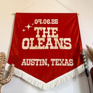 May include: A rust-colored velvet wall banner featuring cream-colored text that reads 04.06.25, THE OLEANS, and AUSTIN, TEXAS. The banner has a pointed bottom edge finished with white fringe and is suspended from a wooden dowel with a white cord.