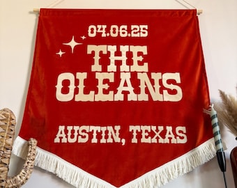 May include: A rust-colored velvet wall banner featuring cream-colored text that reads 04.06.25, THE OLEANS, and AUSTIN, TEXAS. The banner has a pointed bottom edge finished with white fringe and is suspended from a wooden dowel with a white cord.