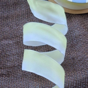 May include: A close-up of a white satin ribbon with a subtle green ombre effect. The ribbon is folded and arranged on a brown burlap surface.
