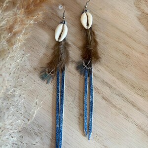 Puede incluir: Un par de pendientes hechos a mano con conchas blancas, plumas marrones y largas tiras de cuero azul denim. Los pendientes tienen ganchos plateados y se exhiben sobre una superficie de madera.