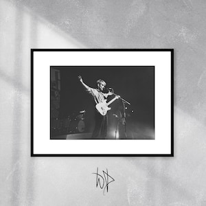 May include: Black and white photograph of a musician performing on stage with a guitar and microphone. The musician is wearing a light-coloured shirt and dark trousers. The stage is lit with spotlights.