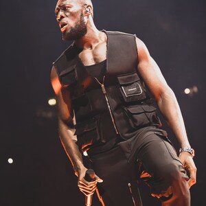 May include: A man wearing a black vest with multiple pockets and black pants, holding a microphone. He is performing on stage in front of a dark background with lights.