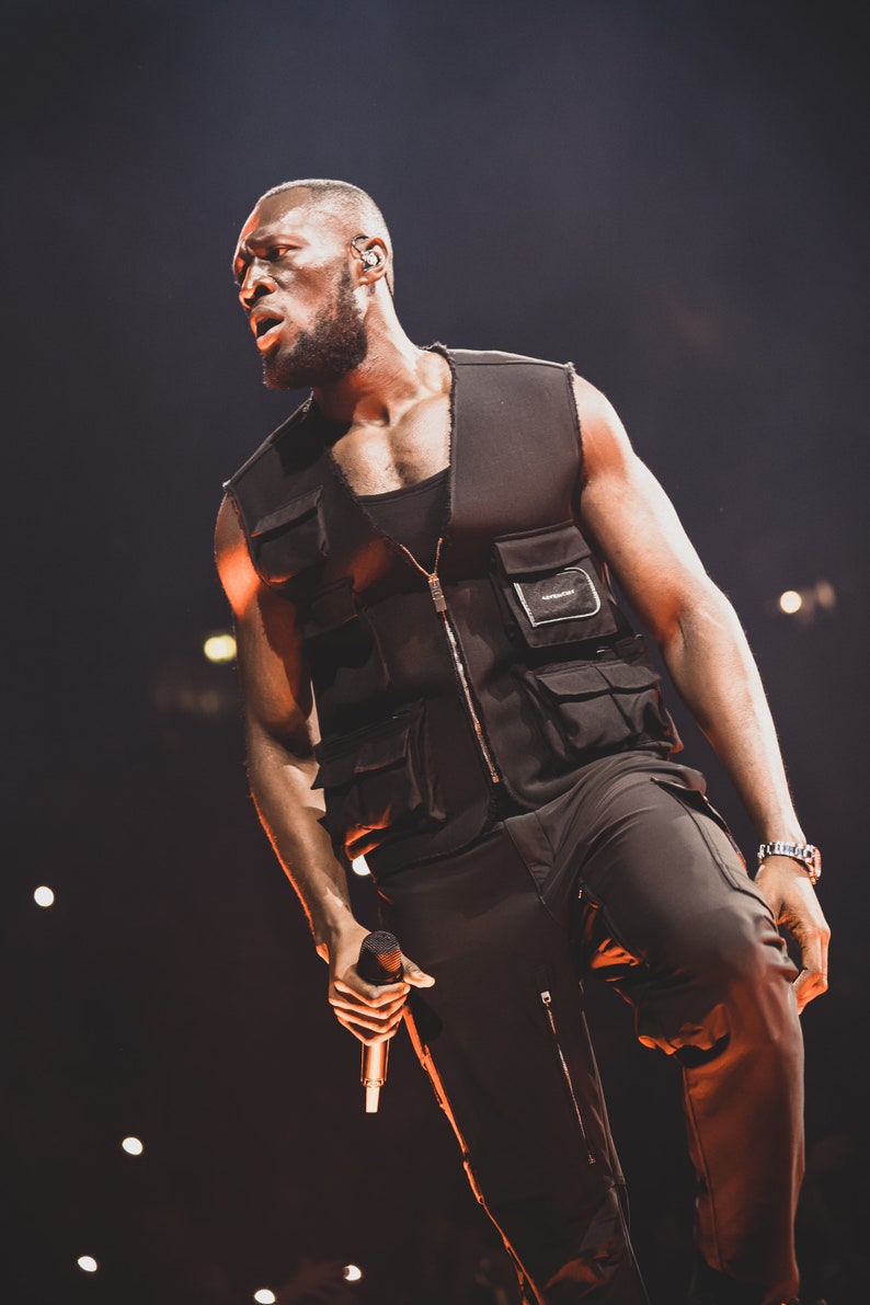 May include: A man wearing a black vest with multiple pockets and black pants, holding a microphone. He is performing on stage in front of a dark background with lights.
