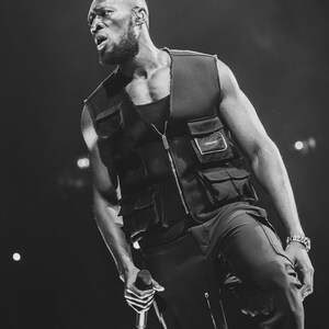 May include: A black and white photo of a person wearing a black vest with a zipper and multiple pockets. The person is holding a microphone and singing into it.