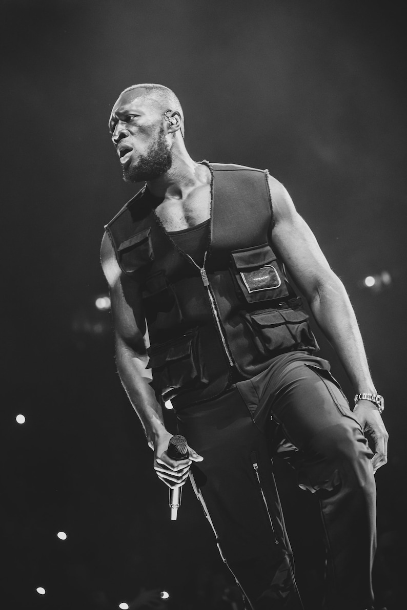 May include: A black and white photo of a person wearing a black vest with a zipper and multiple pockets. The person is holding a microphone and singing into it.