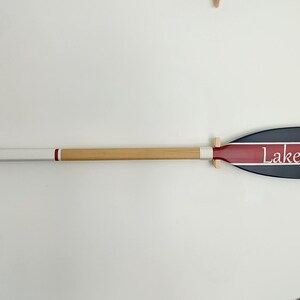 May include: A wooden oar with a blue and red painted blade. The blade has a white stripe and the text "Lake Muskoka" written in white.