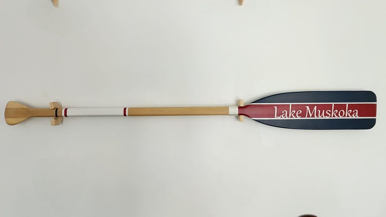 May include: A wooden oar with a blue and red painted blade. The blade has a white stripe and the text "Lake Muskoka" written in white.