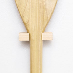 May include: A wooden paddle with a light brown finish is mounted on a white wall using two small wooden blocks.